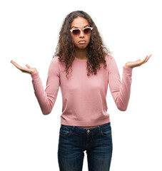 Beautiful young hispanic woman wearing sunglasses clueless and confused expression with arms and...
