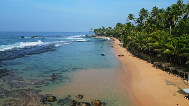 Aerial view of Dalawella beach with turquoise waters, tourists swimming, palm trees lining shore, sandy beach, coastal vibes in Sri Lanka. Ideal for ads, travel vlogs, tourism promos, nature docs.