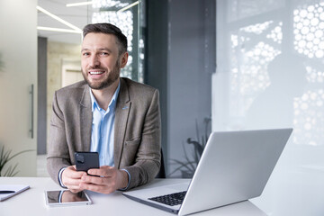 Smiling businessman with smartphone at modern office desk, laptop and tablet visible, expressing job satisfaction and success.