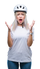 Young blonde woman wearing cyclist security helmet over isolated background crazy and mad shouting and yelling with aggressive expression and arms raised. Frustration concept.