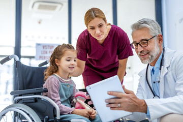 Friendly pediatrician showing someting on tablet to little patient in wheelchair. Cute preschool...