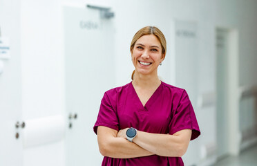 Portrait of beautiful nurse in uniform, standing in the hospital corridor with arms crossed, looking at the camera and smiling.