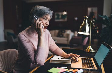 Focused mature woman talking on smartphone and working on laptop