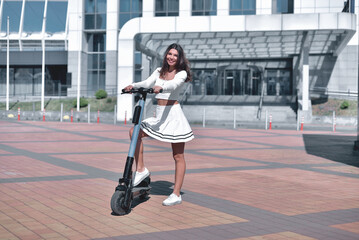Smiling young adult woman posing on an electric scooter parked in front of a modern building