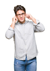 Young handsome man wearing glasses over isolated background covering ears with fingers with annoyed expression for the noise of loud music. Deaf concept.