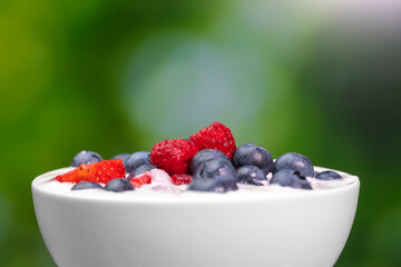 Close-up of a bowl with natural white yogurt and fresh raspberries, blueberries, strawberries on a green blurred background, macro photography. Delicious healthy breakfast