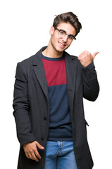 Young handsome elegant man wearing glasses over isolated background smiling with happy face looking and pointing to the side with thumb up.