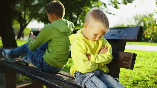 Younger brother sulks as his older sibling is absorbed in smartphone in sunny park. Digital age's impact on childhood interactions. No compromising, not talking. Rivalry concept