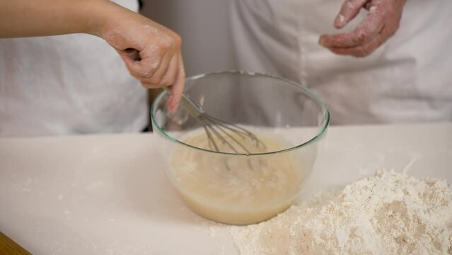 Close up hands little girl mixing yeast dough mixture in bowl. Granny teach granddaughter to cook enjoy process together in cozy kitchen. Home hobby. Older gen share skill with younger generation
