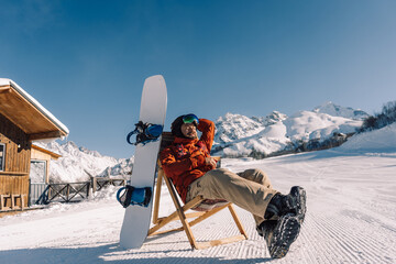 snowboarder sitting in sun lounger on sunny winter day, corduroy of prepared downhill slope at ski...