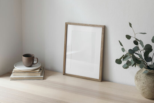 Breakfast still life. Cup of coffee, books. Empty wooden picture frame mockup on desk, table. Textured vase with silver eucalyptus branches. Elegant Scandi working space, home office. Side view.