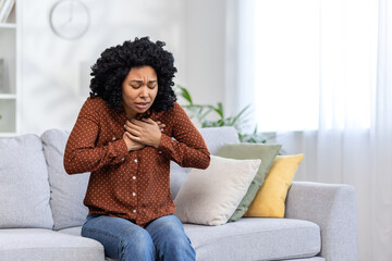 Worried young woman sitting on couch at home feeling chest pain, possible heart attack, stress or...