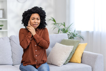 Worried young woman feeling anxious sitting on couch at home with hand on cheek, concept of stress...