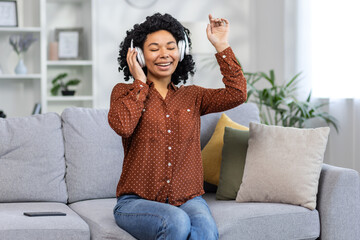 Joyful young woman with headphones enjoying music, dancing on sofa at home in a cozy living room...