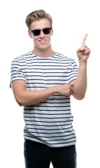 Young handsome blond man wearing sunglasess very happy pointing with hand and finger to the side