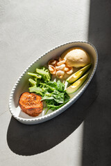 White plate with green salad of baked sweet potato, avacado, spinach, broccoli and shrimp sunlit on gray background