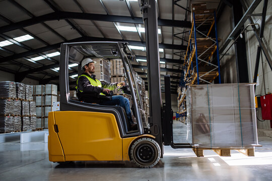 Side view of forklift in warehouse with male driver. Warehouse worker preparing products for shipmennt, delivery, checking stock in warehouse.