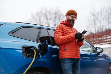 Man charging electric car during cold snowy day, using electric vehicle charging app, checking...