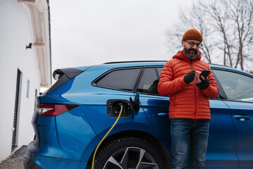 Man charging electric car during cold snowy day, using electric vehicle charging app, checking battery life, energy consumption on smart phone. Charging and driving electric vehicles during winter - obrazy, fototapety, plakaty