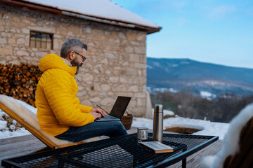 Mature man working from cozy cabin in mountains, sitting on terrace with laptop, enjoying cup of coffee. Concept of remote work from beautiful, peaceful location. Hygge at work. - obrazy, fototapety, plakaty