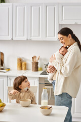Obraz na płótnie Canvas joyful attractive mother posing next to her toddler son with newborn in hands during breakfast