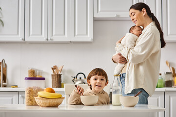 joyous attractive mother posing next to her toddler son with newborn in hands during breakfast