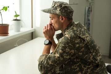 Thoughtful military man staring aside, holding palms by mouth, sitting on couch at home. Young...