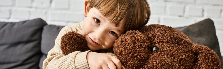 happy little boy hugging his teddy bear while sitting on couch and looking at camera, banner