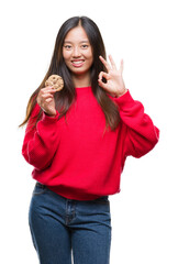 Young asian woman eating chocolate chip cookie over isolated background doing ok sign with fingers, excellent symbol