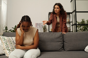 Young irritated brunette woman blaming her upset African American girlfriend sitting on couch and touching temple during quarrel - obrazy, fototapety, plakaty