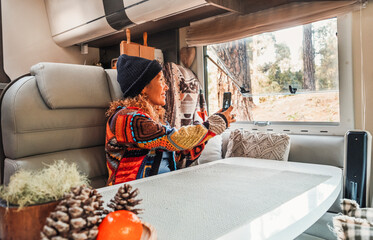 Woman wearing hat and pacwork sweater sitting in camper taking photos with cell phone looking out...