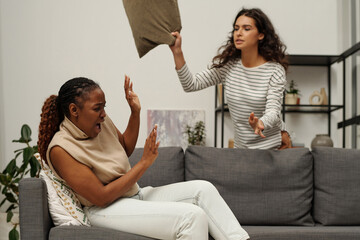 Young angry woman trying to hit her girlfriend with pillow during argument while standing by couch...