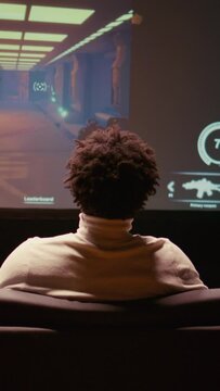 Vertical video BIPOC man playing shooter videogame on ultrawide smart TV in futuristic industrial setting with health, stamina, ammo bars HUD overlay. Zoom in shot of gamer enjoying PvP fps online
