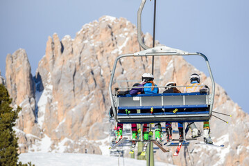 Skiers riding a chairlift in a mountain resort in winter. A snowy rocky peak is in background.