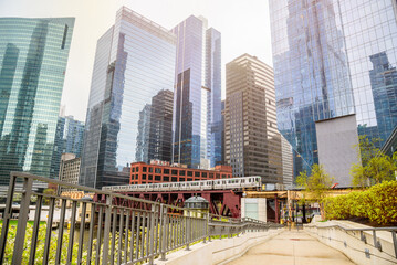 Deserted riverbank path in downtown Chicago with modern galass office towers in background. An elevated train is crossing the river on a double deck bridge.