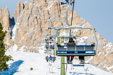 Skiers on a chairlift in a skiing resort in the Alps on a sunny winter day. A towering rocky peak is in background.