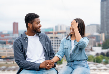 A handsome man and a beautiful woman sitting back to back on the barrier in front of a modern city background dreaming about the future - obrazy, fototapety, plakaty