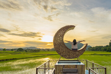Woman sit on crescent moon chair made of rattan for relaxation on bridge in paddy field with...