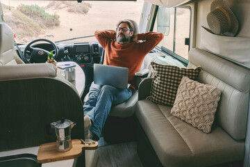 Digital nomad new modern job lifestyle with handsome adult man working and relaxing inside a camper van with beach and nature outside. Smart working free office concept eith laptop and connection - obrazy, fototapety, plakaty