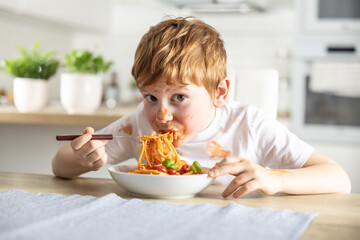 A cute little boy is eating spaghetti bolognese for lunch in the kitchen at home and is covered in...