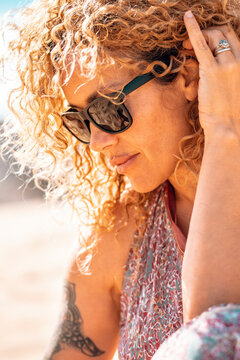 Middle age beautiful woman with curly hair and sunglasses in outdoor side portrait. Modern cute female people with tattoo relax at the beach under the summer sun. Healthy blonde people life