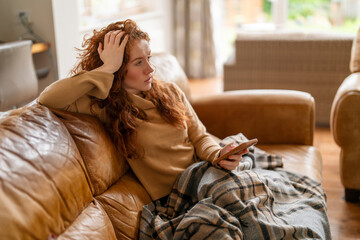 A Beautiful redhead woman watching TV sitting on a sofa at home, watching TV on the couch at home in the living room - obrazy, fototapety, plakaty