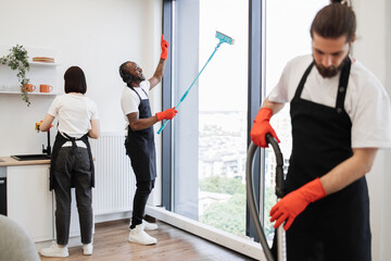 Happy bearded African American cleaning company worker in black apron and headphones washing panoramic windows dancing listening favorite music in bright apartment.