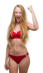 Blonde teenager woman wearing red bikini angry and mad raising fist frustrated and furious while shouting with anger. Rage and aggressive concept.