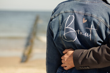 Brautpaar umarmt sich am Strand, Frau trägt personalisierte Jeansjacke mit "Braut"-Aufschrift