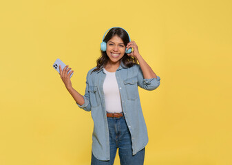 Happy Indian woman carried away with music dancing carefree  wearing  headphones on her ears dressed in jeans and denim shirt, looking at camera, using phone  isolated on  a yellow background