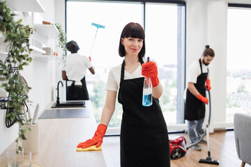 Caucasian janitor in apron and gloves having fun, sprays detergent into camera while cleaning table...