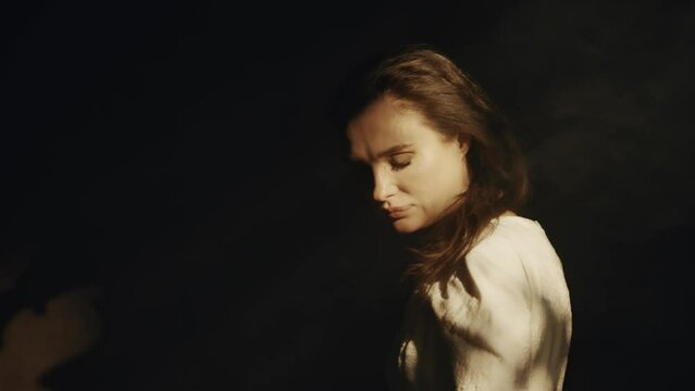 A young woman in a white dress sits in a dark room. Bright sun rays and contrasting shadows. Prayer. Meditation and expression. Fashion and Cinematic concept