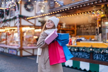 Obraz premium smiling young woman with colorful shopping gift bags having a fun time, using phones, outdoor in an urban winter city. people, communication, shopping and lifestyle concept