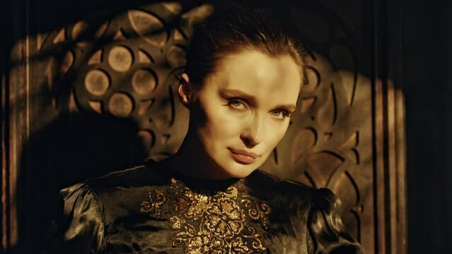 Portrait of a young woman in a black long dress sits in a wooden carved throne in a dark Gothic church. Looks at the camera. Meditation and expression. Fashion and Cinematic concept